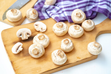 Arrangement of whole and sliced button mushrooms on a wooden cutting board, with a purple cloth and knife in the background. Overhead shot for cooking and kitchen concepts.