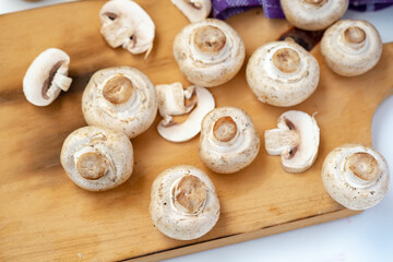 Arrangement of whole and sliced button mushrooms on a wooden cutting board, with a purple cloth and knife in the background. Overhead shot for cooking and kitchen concepts.