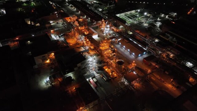 Sprawling industrial facility at night, showing glowing orange structures, steam, and intricate piping systems across a wide manufacturing complex. Aerial drone tilt down.