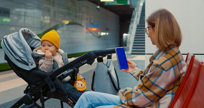 Young mother waiting at subway station, cradling baby in stroller while scrolling smartphone during urban commute, capturing modern parenting moment. Blue screen mockup, chroma key.