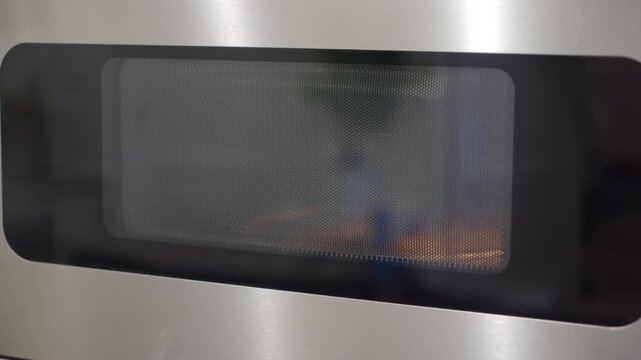 Stainless steel and glass microwave oven. The appliance is heating food, causing a warm glow inside. The result is a steady whirring sound from the cooling fan and the deep hum of the magnetron.