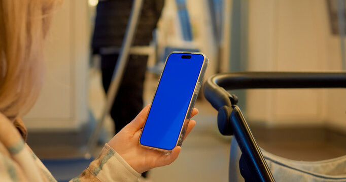 Woman standing on subway platform, holding smartphone with chroma key blue screen while waiting near baby carriage in urban transit environment - Powered by Adobe