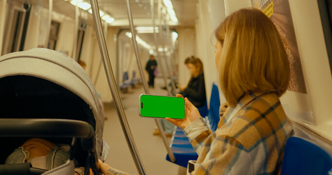 Young mother showing chroma key green screen smartphone while commuting on subway with baby stroller.