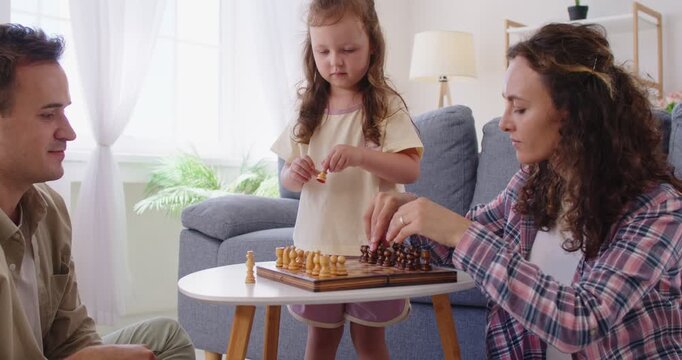 Family with parents and daughter playing chess. Parents smile in living room at home, coaching child girl through strategy on a wooden boardgame. Family leisure and playful learning at home.
