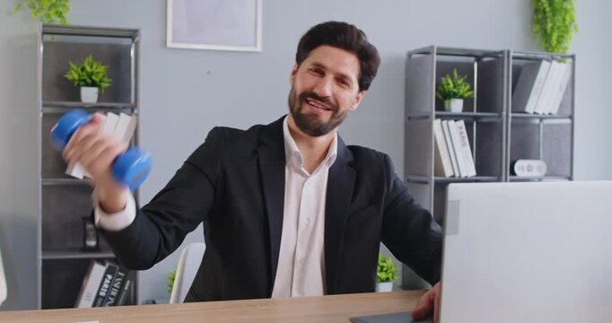 Smiling businessman in suit sitting at desk in office and lifting light weight dumbbell. Cheerful employee doing physical exercises during break, staying active and balancing work with healthy habits.
