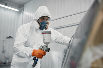 An auto painter dressed in full protective gear is spray-painting a car inside a white garage