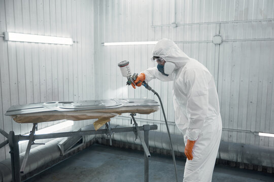 Automotive technician spray-painting car panel in protective suit inside paint booth