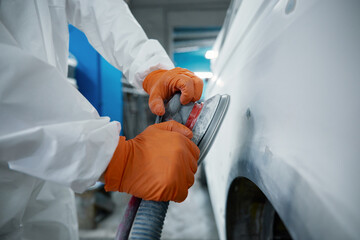 An auto body technician sands a car panel in a protective suit and orange gloves
