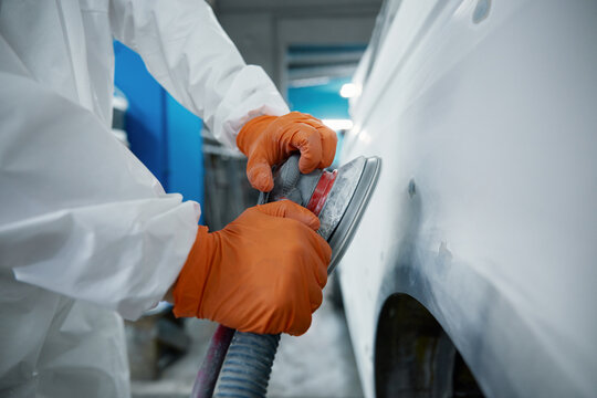 An auto body technician sands a car panel in a protective suit and orange gloves