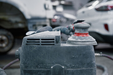 An orbital polisher in a garage shows a close-up of a dusty sander on car bodywork