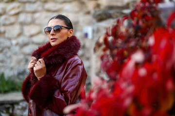 Elegant woman in a burgundy faux-fur coat posing beside vibrant red autumn leaves and stone walls, showcasing fall fashion style in soft natural light.