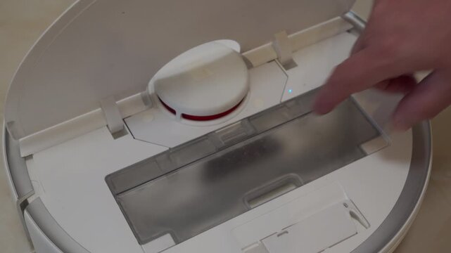 A hand lifts the plastic outer lid on a white robot vacuum cleaner. The action of raising the lid reveals the inner compartment, accompanied by a soft plastic hinge squeaking sound.