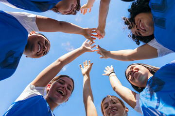 Female soccer players in a huddle, reaching hands together, celebrating team spirit and friendship under blue sky