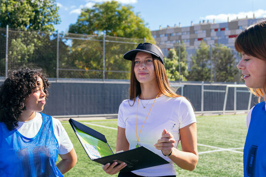 Woman coach holding a tactics board, discussing game strategy with her female soccer team players on a sports field