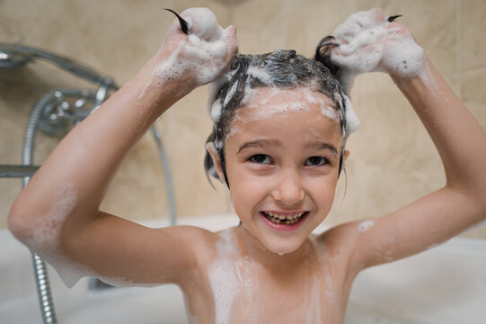 Smiling child washing hair with shampoo foam in bathtub at home
