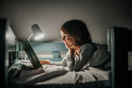 Child reading a book in bed under a lamp at night smiling
