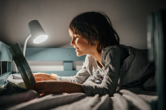 Child reading a book in bed with lamp at night in cozy bedroom