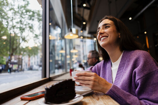 Smiling woman enjoying coffee and chocolate cake at urban cafe window - Powered by Adobe