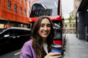 Smiling woman with coffee cup on city street in front of London bus