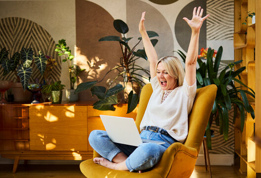 Joyful woman celebrating success while using laptop in sunny home