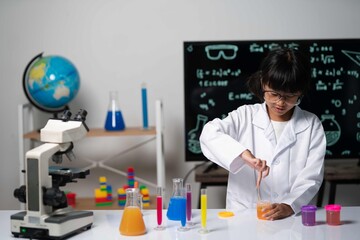 A young girl wearing glasses and a lab coat carefully stirs a bright orange solution. Surrounded by flasks of colorful liquids, she focuses on her chemical mixing process in the science lab.