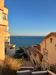 A view down from a hill onto the landscape of a seaside suburb in Spain, bathed in the soft rays of the setting sun. The warm, cozy hues of early autumn are evident. The outlines of white sailboats