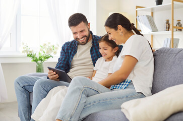 Happy family parents and child at home. Smiling daughter on couch with mother and father using tablet together. Warm living room shows trust, care, and unity. Technology and loving family time.