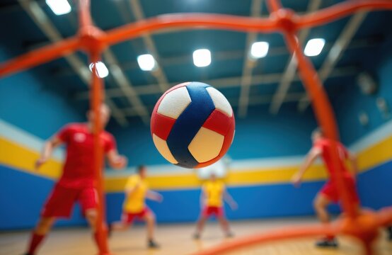 Players in red and yellow uniforms compete in a fast paced indoor ball game. The ball is suspended mid air near the net during intense court action. Team sport competition in gym.