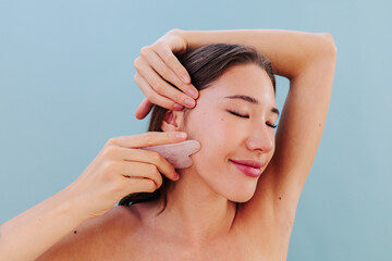 Portrait of woman using gua sha for facial skincare on blue background