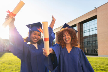 Two university students celebrating graduation outdoors in blue gowns holding diplomas