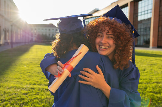 Two university students in cap and gown celebrating graduation outdoors with a joyful embrace