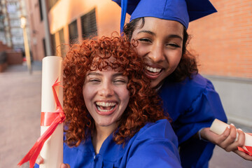 Two young women in blue graduation gowns celebrating their achievement outdoors on a university campus.
