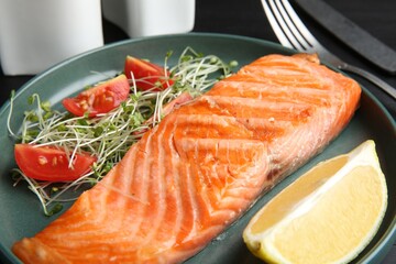 Piece of tasty grilled salmon, lemon, tomato slices and microgreens served on black table, closeup