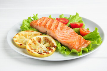 Piece of tasty grilled salmon, lemon slices and salad on white wooden table, closeup
