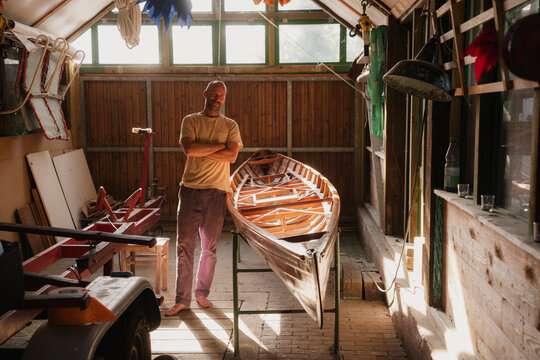 Man standing proudly with wooden boat in sunny workshop