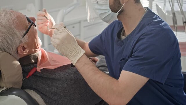 Dentist in protective mask and gloves performing oral examination and treatment on senior patient seated in dental chair in bright contemporary clinic