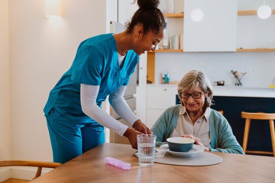 Caregiver serving meal to elderly woman at home kitchen table