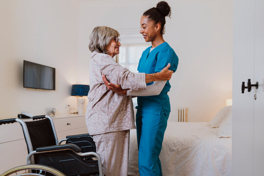 Caregiver helping senior woman transfer from wheelchair to bed at home