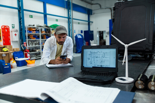 Engineer in a workshop using a phone with a laptop and wind turbine model on the table