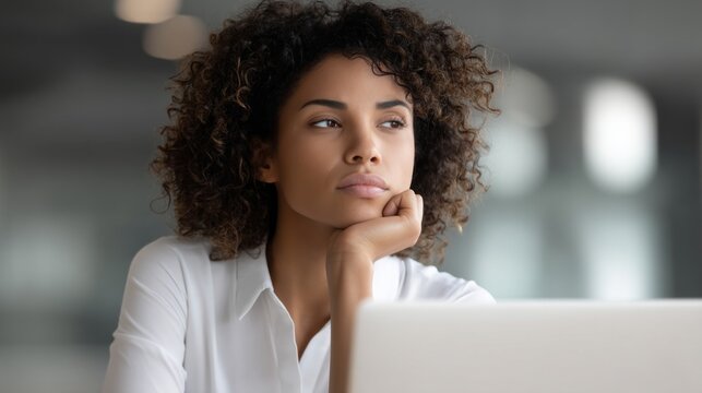 A serious young African American woman sits at a sleek desk, her chin resting on her hand as she gazes thoughtfully at her laptop in a bright office environment
