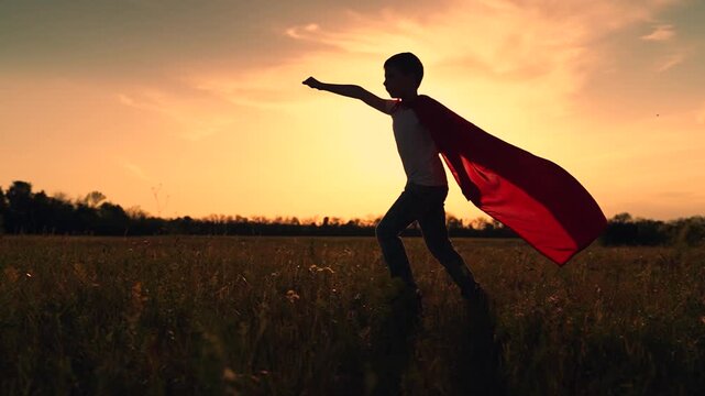Child outdoor superhero play, kid expression emotions. Boy superhero runs across grass park, silhouette. Kid superhero runs in red cape, sunset. Child in superhero costume plays comic book character