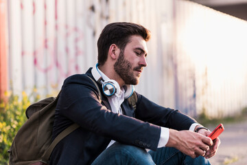 Young businessman using mobile phone and sitting in front of wall at footpath