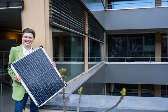 Woman in green jacket holding a solar panel in an office building promoting green energy