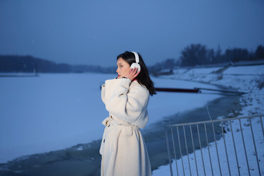 Woman in white coat with headphones by snowy river shore in winter