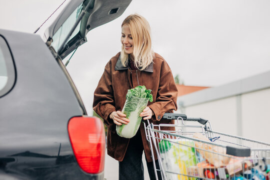 Smiling person loading groceries into car at supermarket parking lot