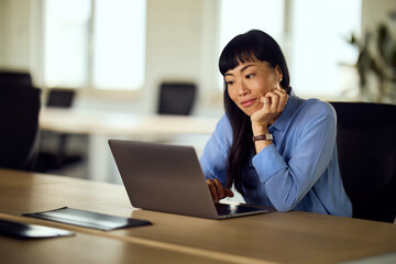 Asian Woman Working on Laptop in Modern Office, Focused and Thoughtful at Desk