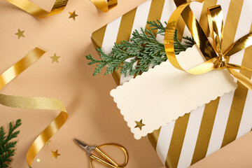 Close-up of gift wrapped in gold and white striped paper with blank tag, and cedar sprig on beige background with items for wrapping. Copy space