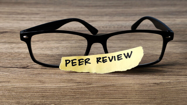 Peer Review concept with a handwritten note placed on reading glasses on a wooden desk, symbolizing academic research and quality assurance.