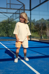 Blond young boy standing on bright blue tennis court with hands in pockets, wearing casual beige oversized t-shirt, orange shorts, and white sneakers, looking serious under clear sunny sky