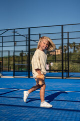 Blond young boy walking confidently across bright blue tennis court with hands in pockets, dressed in beige oversized t-shirt, orange shorts, and white sneakers